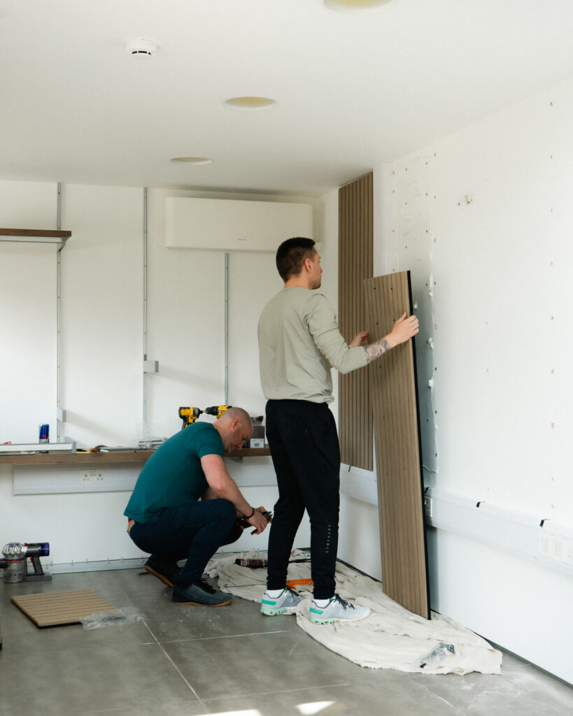 Man crouched looking at wall whilst another man mounts a wall panel to wall.