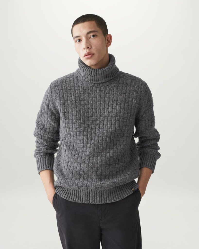 Man standing with his hands in his pocket wearing a grey turtleneck.